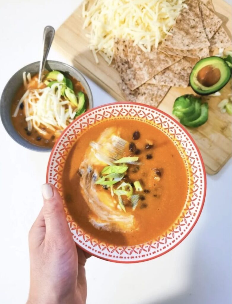 Un bol de soupe rouge avec des morceaux de poisson et des oignons verts, tenu par une main, accompagné d'un petit bol de soupe, de fromage râpé, de tortillas croustillantes et d'avocat sur une planche en bois.
